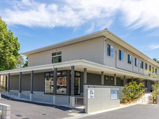 緑に囲まれた2階建ての建物は1階部分がダークグレー、2階部分が白で、複数の介護事業所名が書かれたプレートが門に貼られている。