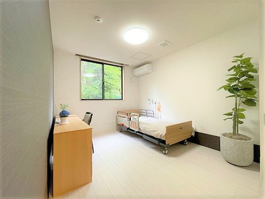寝具がセットされたベッドがのそばにはオレンジの緊急通報装置が設置され、タンスやデスクセット、観葉植物が置かれている。