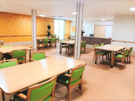 木製のダイニングテーブルが複数置かれ明るい雰囲気。観葉植物もあり、壁には手すりが付いている。