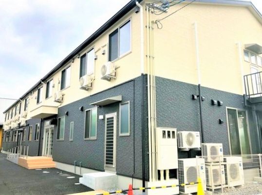 ベージュとグレー2階建ての建物は横に長く、出入り口が複数付き、駐車スペースもある。