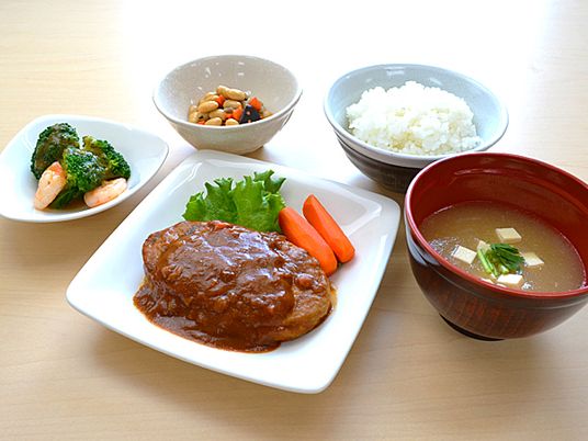 大きなハンバーグにはデミグラスソースがかかり、野菜が添えられている。白いご飯やみそ汁もある。