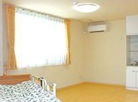 エアコンが設置された広いフローリングの部屋にはキッチンが付き、寝具がセットされたベッドが置かれ、窓にはレースカーテンとカーテンが付いている。
