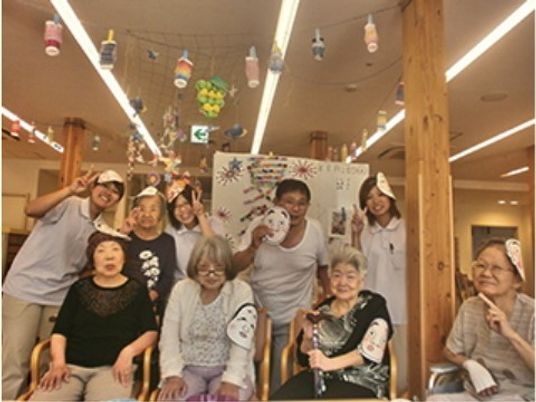 ひょっとこのお面をかぶった入居者とスタッフが、館内の飾りの下で集合写真を撮っている。