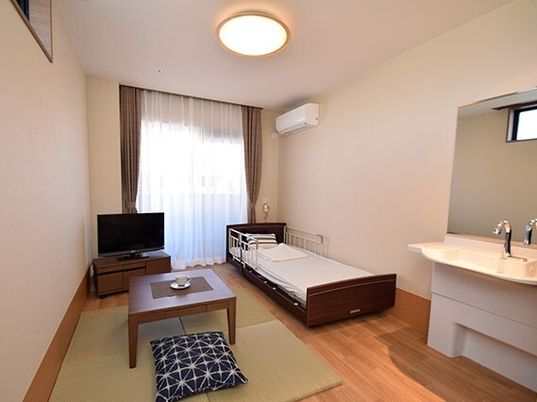 エアコンが付いたフローリングの部屋には寝具付きのベッドやテレビ、車いす対応の洗面所があり、畳風マットの上にはちゃぶ台がある。