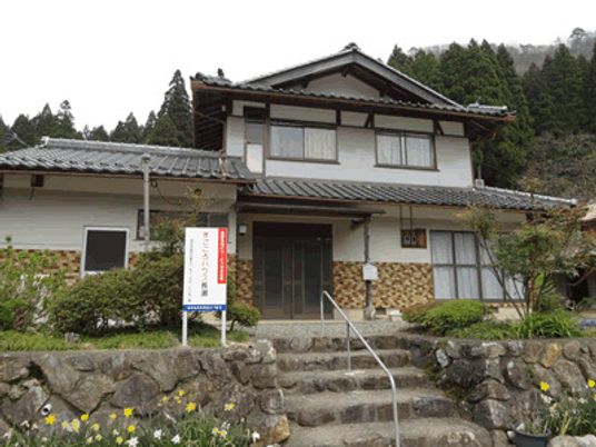 自然に囲まれた緑豊かな場所に建つ2階建ての民家風の住宅。瓦屋根で1階部分よりも2階部分が小さく、玄関の前の階段には手すりが付いている。