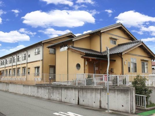２階建ての建物は日当たり良好でブラウン系の外壁。建物の周りにはフェンスが設置されている。