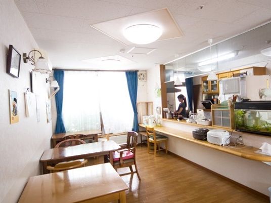 青いカーテンが設置された大きな窓があり日当たり良好。対面式のキッチンには女性スタッフがいる。