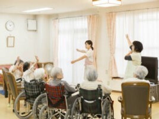 体操をしているエプロンを付けた女性スタッフが2名。スタッフを見ながら車いすやイスに座った高齢女性も体操をしている。
