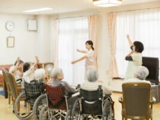 体操をしているエプロンを付けた女性スタッフが2名。スタッフを見ながら車いすやイスに座った高齢女性も体操をしている。