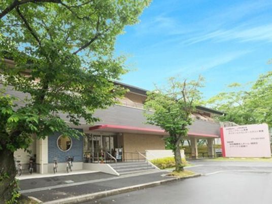 2階建ての建物2階部分に大きなベランダが付いていて、玄関には階段とスロープがあり、広い駐車場内には緑も植えられている。