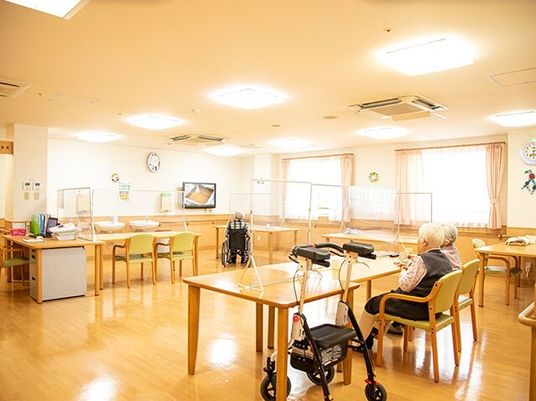 フローリングの食堂にはダイニングテーブルが複数置かれ、大きな窓やテレビがあり、高齢女性2名が話している。