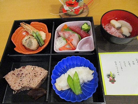 かぶら蒸しや扇形の赤飯、エビなどの煮物、刺身盛り合わせ、吸い物が並んだ黒いトレー。