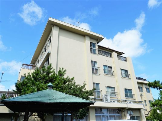 鉄骨造数階建ての建物の外観写真。手前に入り口らしき瓦屋根、奥のほうに建物がある