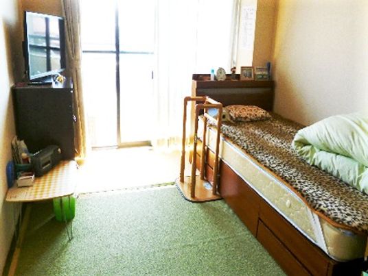 畳の部屋にベッドとテレビなど日用品が置いてある様子。奥のほうに窓が置いてある空間