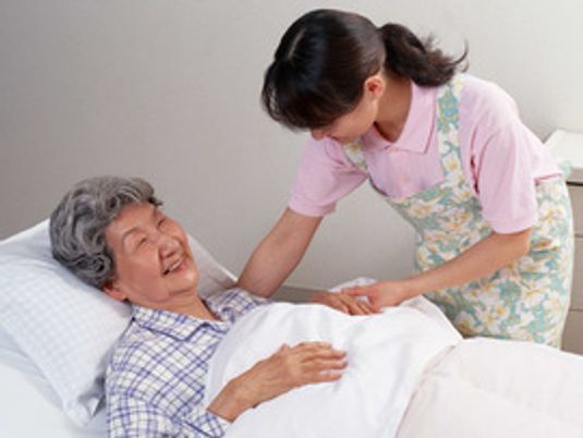 ベッドに横たわっている高齢女性に対して優しく微笑みかける女性スタッフ。