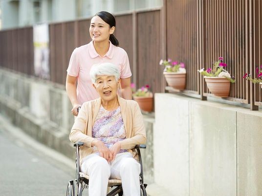 住宅地の中を散歩している車いすの高齢女性と付き添いの若い女性スタッフ。スタッフはポロシャツを来て、微笑みながら車いすを押している。