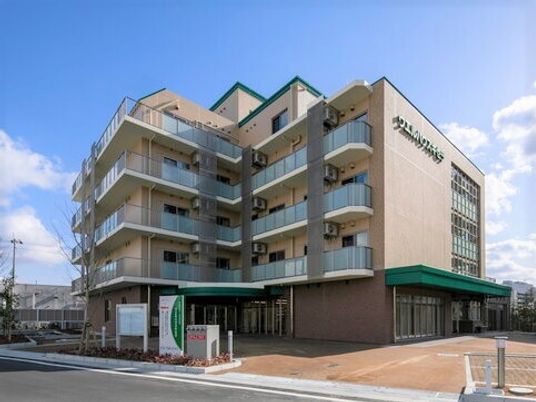 7階建てのベージュの建物はエル字型で、各フロアにベランダが付き、外壁にはホーム名が書かれている。