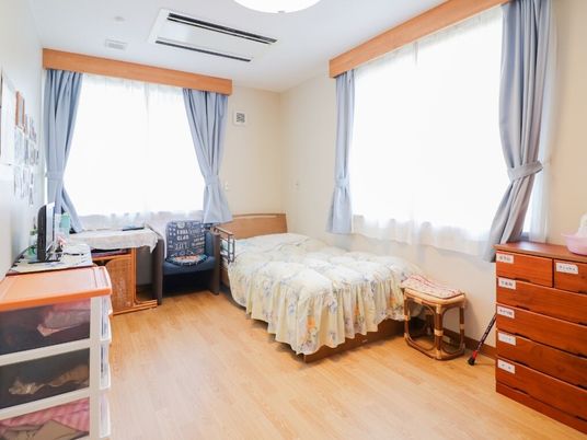 寝具付きのベッドやタンス、いすや収納ケースが置かれたフローリングの部屋には大きな窓が二つ付き、カーテンとレースカーテンが付いている。