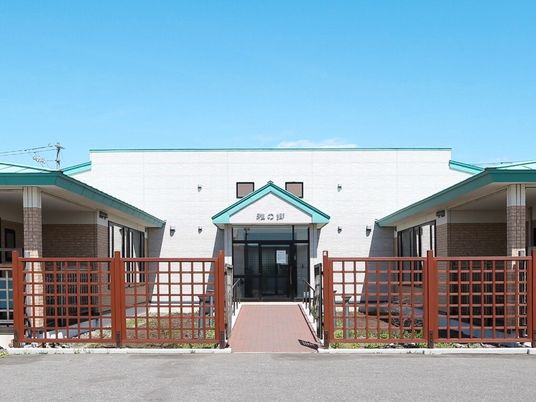 白い外壁の平屋建ての建物の玄関には緑の屋根が付いていて、駐車場から玄関までまっすぐな道が伸びている。周りにはフェンスもある。