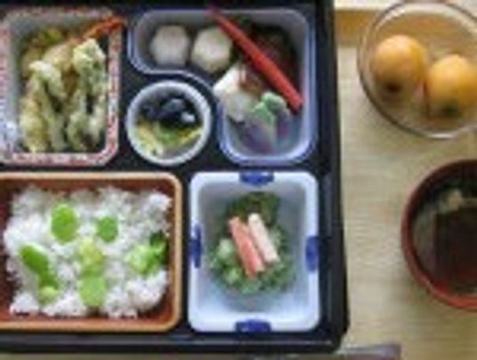 豆ごはんや天ぷらが詰められたお弁当風の食事の横にはビワの実やみそ汁がある。