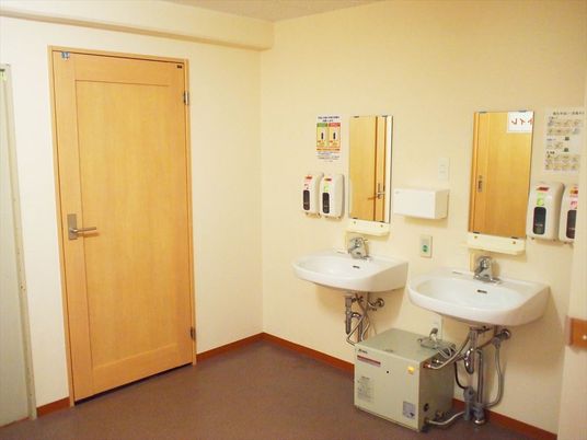 左側に洗面所が二つ並び、それぞれの鏡や壁掛け式のソープなどがある。左側に木目調のドアがある。
