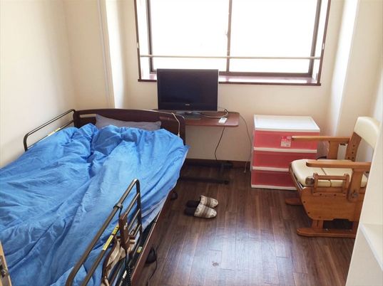 シンプルな居室には、入居者が持ち込んだテレビや家具調ポータブルトイレ、収納ケース、そして介護用ベッドがある。