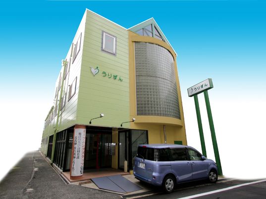 ライトグリーンとイエローの3階建ての建物。外壁にはホーム名が書かれていて、玄関前には一台の車が止まっている。