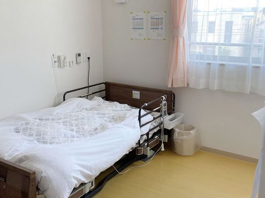 寝具付きのベッドのそばには緊急通報装置があり、大きな窓にはレースカーテンとカーテンが付いている。