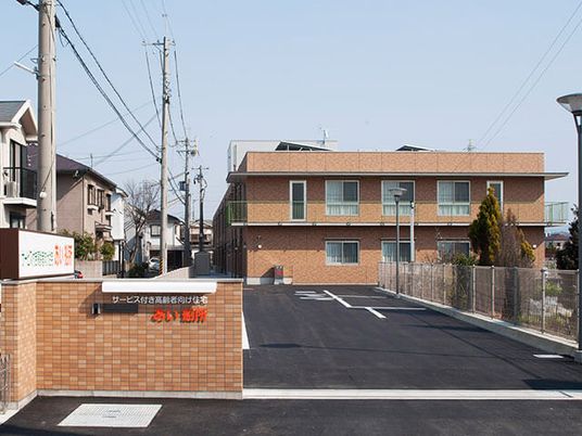 広い駐車場もあり、落ち着いた雰囲気の2階建ての建物。ブラウン系で門には赤い文字で住宅名が書かれている。