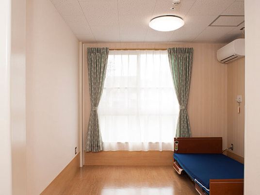 介護ベッドやエアコン、カーテン付きの大きな窓があるフローリングの居室。壁には緊急通報装置を設置している。
