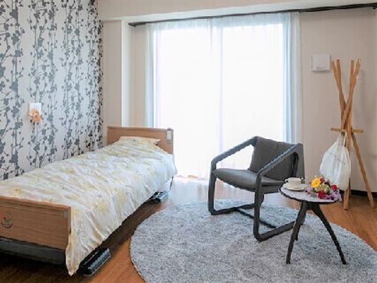 寝具付きのベッドが置かれたフローリングの居室にはテーブルセットやハンガーラックなどの家具やベッドもあり、大きな窓が付き明るい雰囲気。