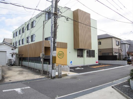 木のあしらいがある鉄骨造の建物の様子。手前に駐車場スペースがあるホームの外観