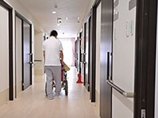 廊下を車いすで通っていくスタッフと高齢者の後ろ姿。両側に個室のドアが見えている写真
