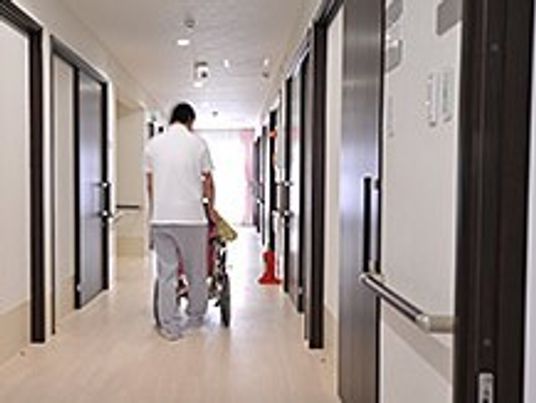 廊下を車いすで通っていくスタッフと高齢者の後ろ姿。両側に個室のドアが見えている写真
