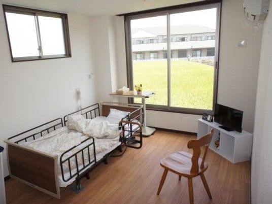 窓の外には自然が広がっている。居室内にはベッドやテレビなどの家具家電が置かれ、温かみのある雰囲気。