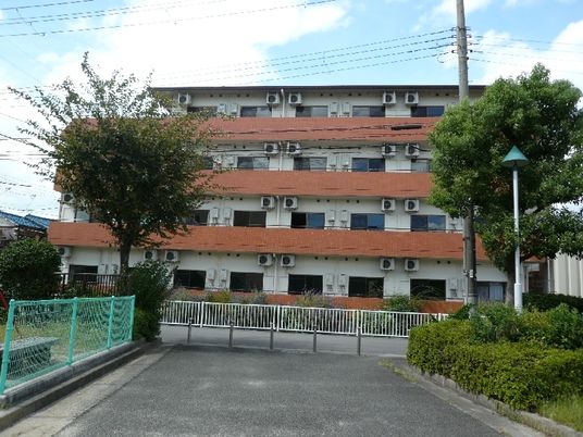 角フロアにベランダが付いた4階建ての建物の周りには草木が植えられ、外壁は白とオレンジ。