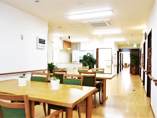 木製のダイニングテーブルが複数置かれ、キッチンはオープンで、右側には居室が並び、明るい雰囲気。