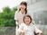 屋外で笑顔で過ごしている車いすの高齢女性と付き添いのピンクのエプロンを付けた若い女性スタッフ。