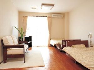 ベッドが二つ置かれ、そばには緊急通報装置が設置され、ソファやテレビ、エアコンなどもあるフローリングの居室。