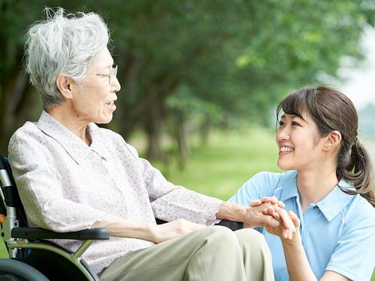 眼鏡をかけた高齢女性が車いすに座り、緑あふれる屋外で外気浴を楽しんでいる。そばにいる女性スタッフは腰を下ろし、女性の手を握っている。