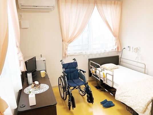 レースカーテンとカーテンが付いた大きな窓が二つある部屋には寝具付きのベッドや車いす、エアコンがある。