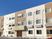 ベージュとブラウンの3階建ての建物。住宅名が書かれた屋根が玄関に付き、敷地内には駐車場もある。