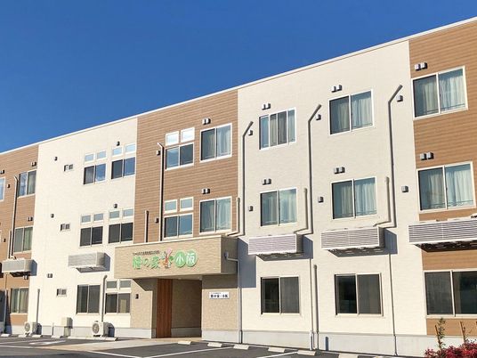 ベージュとブラウンの3階建ての建物。住宅名が書かれた屋根が玄関に付き、敷地内には駐車場もある。