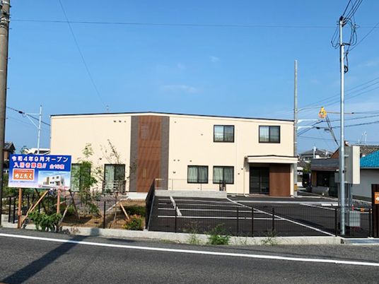 ベージュの外観の２階建ての建物の周りには草木が植えられた庭があり、広い駐車場も完備されている。