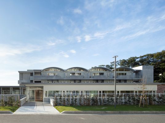 青い空の下に建っている白い建物は横に長く、玄関には白いアプローチが続いていて、芝生スペースもある。建物の周りには緑がある。