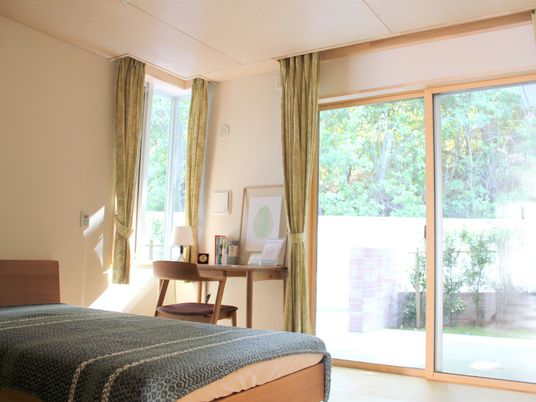 大きな窓がたくさんついたフローリングの部屋は明るい雰囲気。寝具がセットされたベッドが置かれ、コーナーにはデスクセットがあり、ランプも付いている。