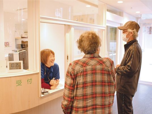 眼鏡をかけて帽子とマスクをした高齢男性と高齢女性が受付の前に建っていて、受付にいる女性スタッフが笑顔で対応している。