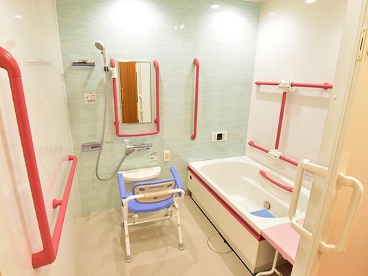 赤い手すりがたくさんついた一人用の浴室。浴槽横にはベンチが設置され、シャワーチェアも置かれている。