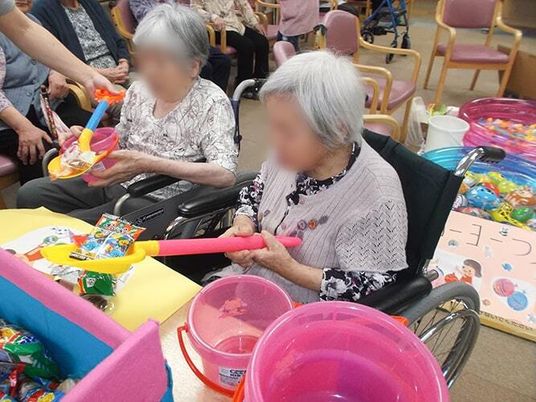 夏祭りをホーム内で行っている様子。手前にはスコップでお菓子をすくっている高齢女性たちがいて、奥にはヨーヨー釣りがある。