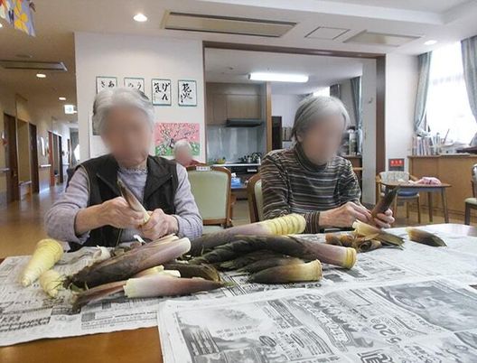 テーブルの上には新聞紙が敷かれ、２名の高齢女性たちがタケノコの皮をむいている。
